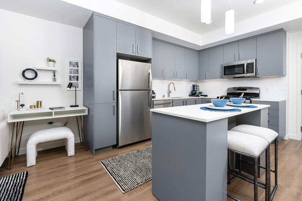 Combined living room and kitchen area with a gray sectional sofa, abstract wall art, and modern kitchen appliances in a bright space.