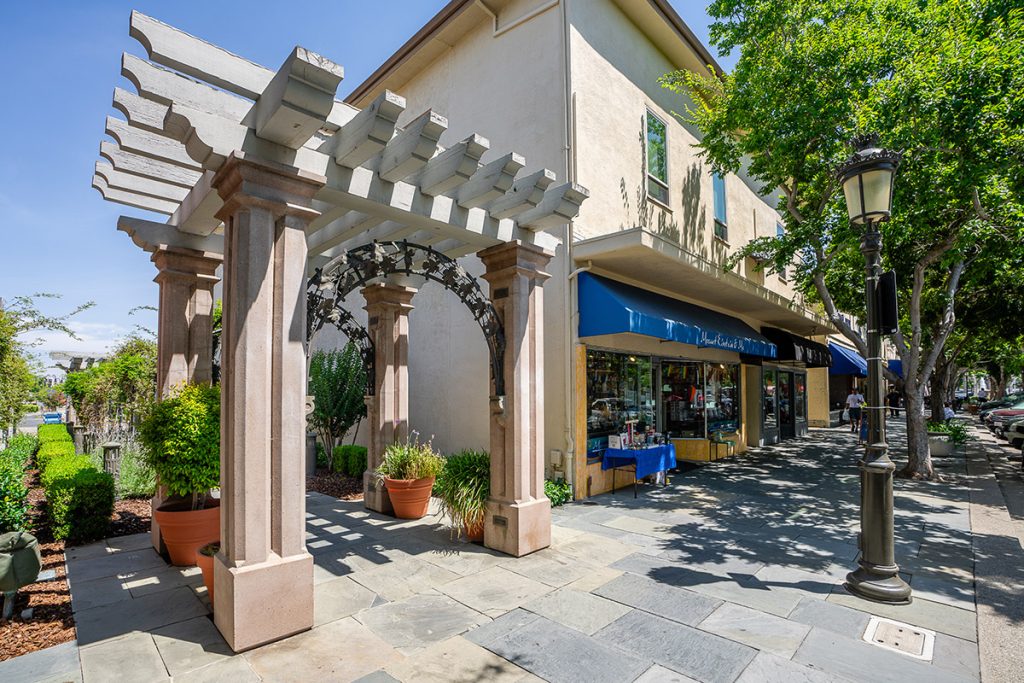 A shaded sidewalk in a quaint shopping district, featuring a pergola with classical columns and an intricate trellis, complementing the storefronts with blue awnings under a bright blue sky