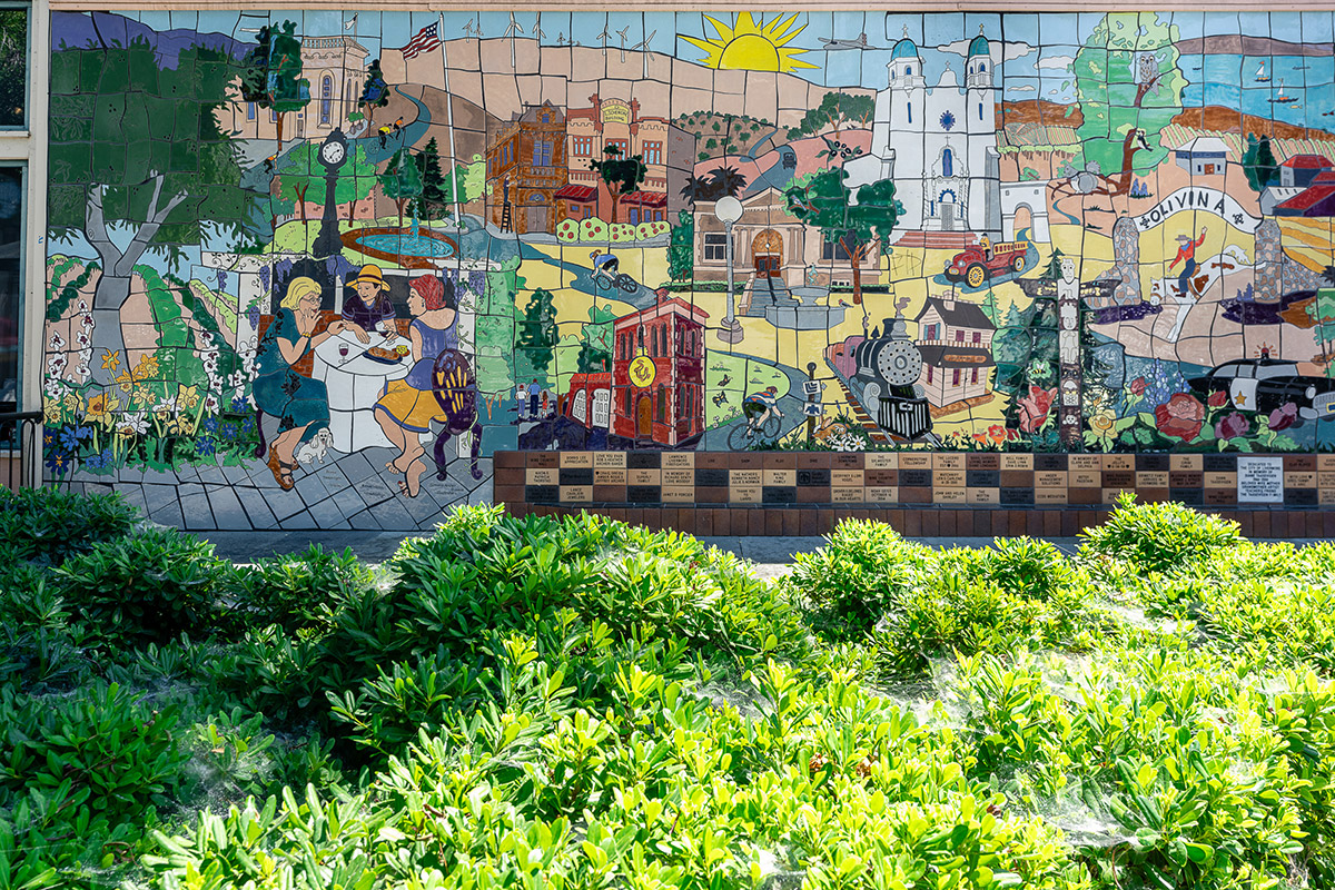 Colorful mosaic mural depicting community scenes, iconic buildings, and nature. Characters dine, cycle, and interact amid landmarks. Foreground with lush green shrubs.