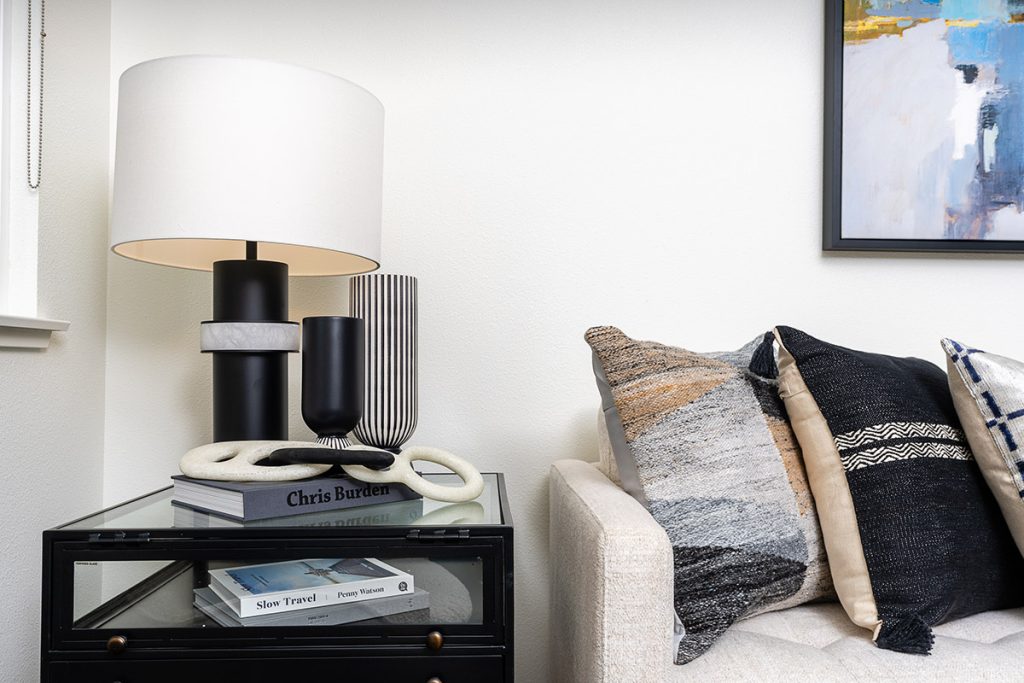 Close-up of a living room corner with a white sofa, eclectic throw pillows, and a white lamp atop books on a glass side table.