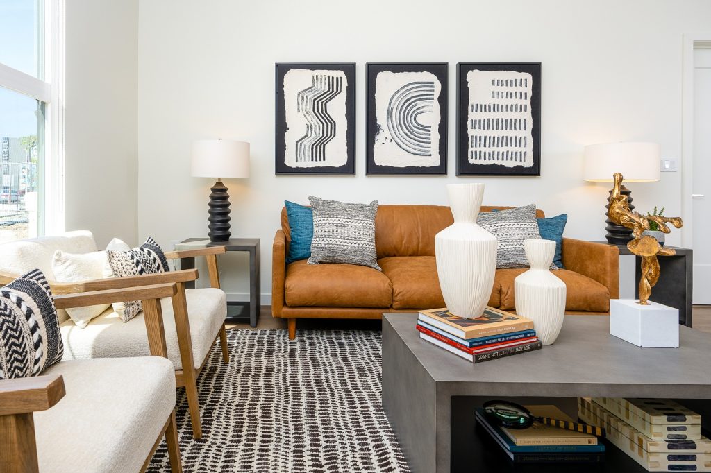 Bright and inviting living room with a plush caramel-colored couch, patterned throw pillows, and black and white artwork, in a modern apartment setting.